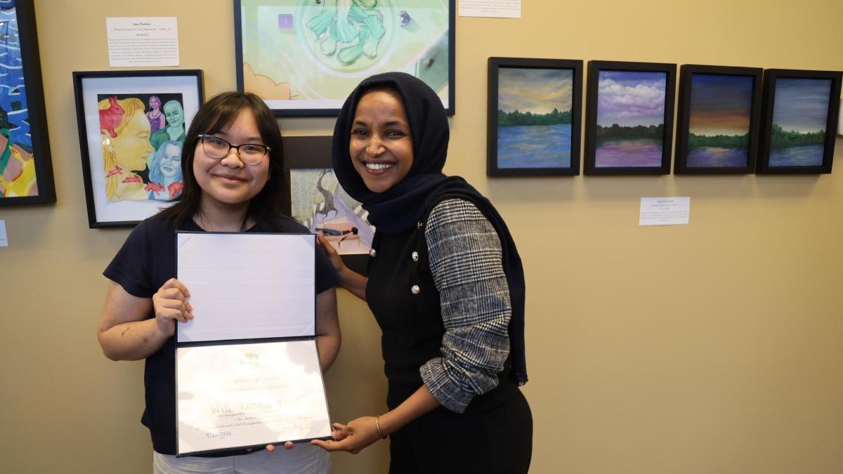 Rep. Omar presents the 2026 Congressional Art Competition winner with a certificate. The two stand in front of the winning artwork, which is framed on the wall behind them.
