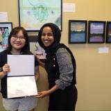 Rep. Omar presents the 2026 Congressional Art Competition winner with a certificate. The two stand in front of the winning artwork, which is framed on the wall behind them.